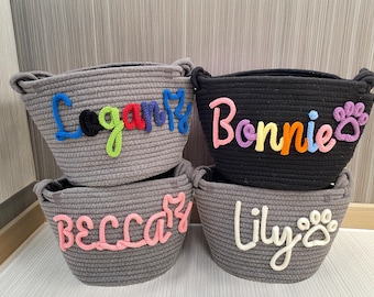 Puede incluir: Cuatro cestas de cuerda en gris y negro, cada una con un nombre bordado en la parte delantera. Los nombres son Logan, Bonnie, Becca y Lily. La cesta de Lily tiene un diseño de huella de pata.