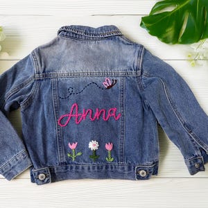 May include: A blue denim jacket with the name "Anna" embroidered in pink script on the back. The jacket features a butterfly and flower embroidery. The jacket is a children's size.