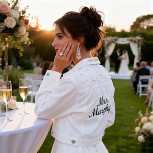 May include: A white denim jacket featuring pearl embellishments on the shoulders and back, with the text Mrs. Murphy embroidered in black script on the back. The jacket has a collar, buttoned cuffs, and a button-front closure, worn over a white garment.