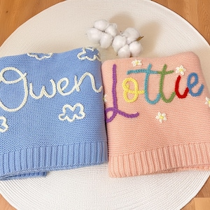 May include: Two personalised baby blankets. One is light blue with the name "Owen" in white script and cloud accents. The other is pink with the name "Lottie" in rainbow script and flower accents. Both are knitted.