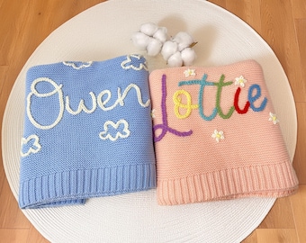 May include: Two folded knitted baby blankets. One is light blue with the name "Owen" in white script and white cloud designs. The other is pink with the name "Lottie" in rainbow colours and small white flower designs.