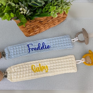 May include: Two baby pacifier clips with waffle-textured fabric. One is light blue with "Freddie" embroidered in blue, the other is cream with "Baby" in yellow. Each has a clip and a pacifier.