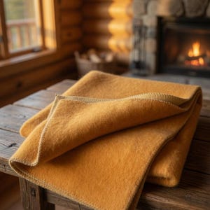 Puede incluir: Una manta amarilla mostaza doblada, con un borde cosido de color claro, descansa sobre una mesa de madera rústica. La textura de la manta parece suave y acogedora. El fondo presenta una chimenea acogedora y una ventana.