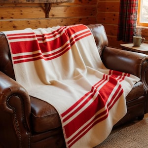 Puede incluir: Manta color crema con llamativas rayas rojas, sobre un sillón de cuero marrón. La manta presenta un diseño clásico con gruesas bandas rojas y rayas blancas más finas. El acogedor plaid añade un toque de calidez y encanto rústico al espacio.