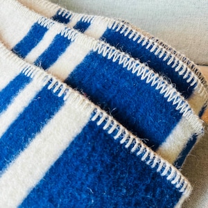 Op de afbeelding: Een gevouwen deken met een patroon van dikke, opvallende blauwe strepen die afwisselen met gebroken witte strepen. De randen van de deken zijn afgewerkt met een zichtbaar, dik festonsteekdetail in crèmekleurig garen, wat de textuur benadrukt.