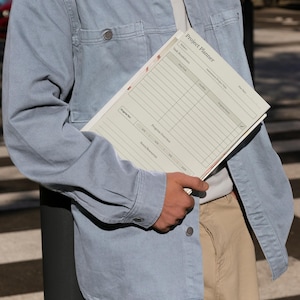 May include: A person holding a Project Planner. The planner is light-colored with a grid layout and the title "Project Planner" at the top. The person is wearing a light blue denim shirt and khaki pants.