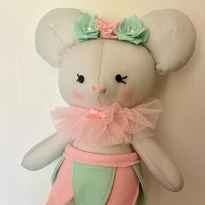 Könnte beinhalten: Eine handgefertigte Plüschmauspuppe mit großen Ohren, die ein rosa-grünes Outfit trägt. Die Puppe hat einen rosa Tüllkragen, einen Blumenkranz und grüne Filzschuhe. Die Puppe steht auf einer weißen Basis.