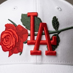 Puede incluir: Gorra de béisbol blanca con una rosa roja bordada y las letras "LA" en rojo. La gorra tiene una visera curva y un diseño clásico de seis paneles. La rosa y las letras están detalladas con hojas verdes.