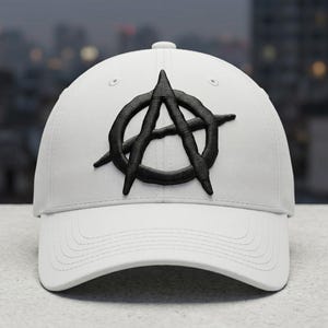May include: White baseball cap with a black embroidered anarchy symbol on the front. The cap has a curved brim and a button on top. The background is blurred, suggesting an outdoor setting.
