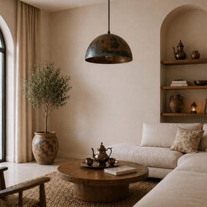 K&ouml;nnte beinhalten: Ein Wohnzimmer im rustikalen Stil mit einer gro&szlig;en, kuppelf&ouml;rmigen Pendelleuchte mit verwitterter Kupferoberfl&auml;che. Der Raum umfasst einen runden Couchtisch aus Holz, ein wei&szlig;es Ecksofa und einen Olivenbaum im Topf, was eine warme Atmosph&auml;re schafft.