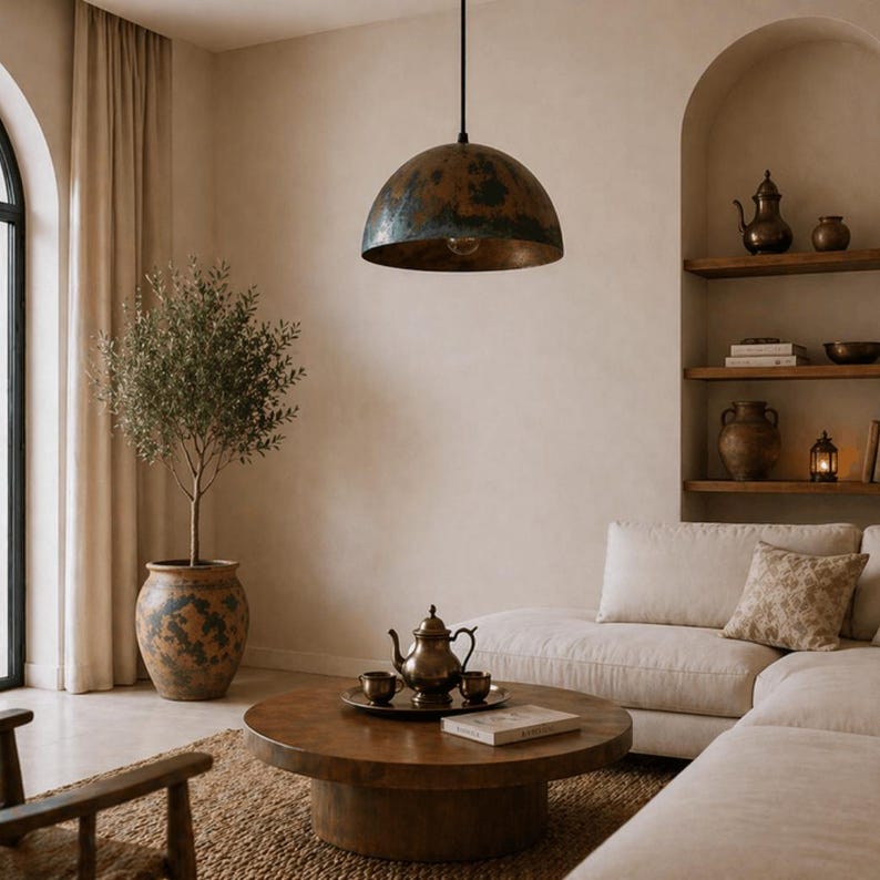 K&ouml;nnte beinhalten: Ein Wohnzimmer im rustikalen Stil mit einer gro&szlig;en, kuppelf&ouml;rmigen Pendelleuchte mit verwitterter Kupferoberfl&auml;che. Der Raum umfasst einen runden Couchtisch aus Holz, ein wei&szlig;es Ecksofa und einen Olivenbaum im Topf, was eine warme Atmosph&auml;re schafft.