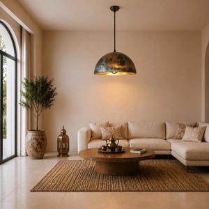 K&ouml;nnte beinhalten: Ein Wohnzimmer mit einer gro&szlig;en, gealterten Metall-Pendelleuchte &uuml;ber einem runden Couchtisch aus Holz. Ein beiges Ecksofa mit gemusterten Kissen befindet sich gegen&uuml;ber einem gro&szlig;en Fenster. Ein Jute-Teppich und eine Topfpflanze vervollst&auml;ndigen den warmen, einladenden Raum.