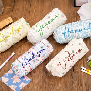 Puede incluir: Cinco estuches cilíndricos para lápices con texturas de tela acolchada, cada uno con un patrón floral o de cuadros único y un nombre personalizado bordado en letra cursiva: Amelia, Gianna, Hannah, Madison y Victoria, con cremalleras visibles.