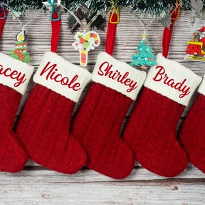 Puede incluir: Cinco calcetines navideños rojos con puños blancos, cada uno bordado con un nombre en escritura roja. Adornos decorativos cuelgan sobre los calcetines. Los calcetines se muestran sobre un fondo de madera.