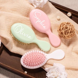 May include: Three oval hairbrushes with handles. One is light green with the name Olivia in white script, one is light pink with the name Sophia in white script, and one is white with pink bristles and a pink cushion base.