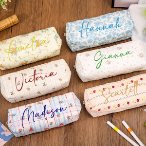 Puede incluir: Seis estuches rectangulares acolchados con estampados florales y varios diseños de cuadros. Cada estuche está personalizado con nombres bordados, incluidos Amelia, Hannah, Gianna, Victoria, Scarlett y Madison, en diferentes fuentes cursivas.