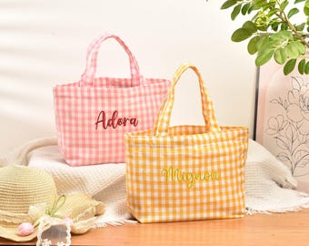 May include: Two small tote bags made of gingham fabric. One bag is pink and white with the name Adora embroidered in dark pink. The other bag is yellow and white with the name Mignon embroidered in yellow. Both bags feature two top handles.