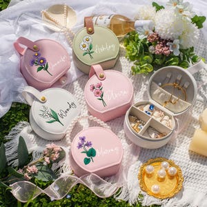 Puede incluir: Una colección de joyeros redondos y personalizados en varios colores, incluyendo rosa, blanco y verde. Cada caja presenta un diseño floral y un nombre. Una caja está abierta, revelando compartimentos llenos de joyas. Una bandeja dorada contiene perlas.