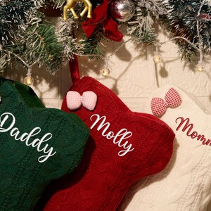 May include: Three cable-knit Christmas stockings in dark green, red, and cream. The green stocking features the white embroidered name Daddy, the red stocking features the white name Molly, and the cream stocking features the red name Mommy. Each stocking is adorned with a small gingham bow.
