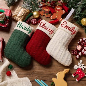May include: Three cable-knit Christmas stockings in dark green, deep red, and cream. Each stocking features a white cuff embroidered with the names Hardy, Stacey, and Emily in green and red script, arranged horizontally on a wooden surface.