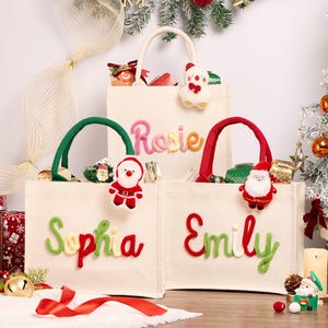 Pode incluir: Três sacolas de lona na cor off-white com enfeites de pelúcia do Papai Noel presos às alças. Cada bolsa exibe um nome em letras cursivas coloridas e em relevo: Rosie, Sophia e Emily. As bolsas possuem alças de tecido nas cores vermelha ou verde.