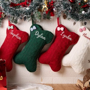 May include: Four bone-shaped Christmas stockings in red, green, and white cable knit fabric. Each stocking features a small gingham bow at the top and is personalized with the embroidered names Sophia, Dylan, Lyuba, and Hannah in white or red script.