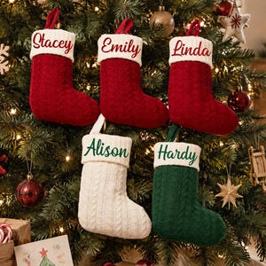 May include: Five cable-knit Christmas stockings with white cuffs. Three red stockings feature the names Stacey, Emily, and Linda in red script. One white stocking features the name Alison in green script, and one green stocking features the name Hardy in green script.