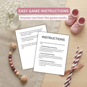 May include: Two white paper sheets with black printed text titled INSTRUCTIONS for a baby shower bingo game. The sheets are surrounded by a glass vase with white flowers, two small pink baby shoes, a wooden bead pacifier clip, and striped straws.