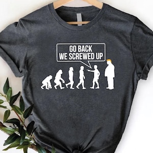 May include: Dark gray t-shirt with a humorous graphic depicting the evolution of man, culminating in a figure with orange hair. The text "GO BACK WE SCREWED UP" is above the figures. The shirt is displayed on a wooden hanger.
