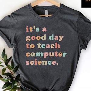 May include: Dark gray t-shirt with the text "it's a good day to teach computer science" in colorful letters. The shirt is hanging on a wooden hanger. A sprig of greenery is placed on the bottom left of the shirt.