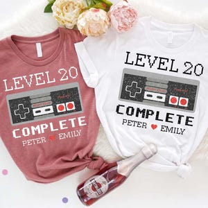 May include: Two t-shirts, one mauve and one white, with a retro video game controller graphic. The text on the shirts reads "LEVEL 20 COMPLETE" with the names "Peter" and "Emily" and a heart symbol. A bottle of champagne is in the foreground.