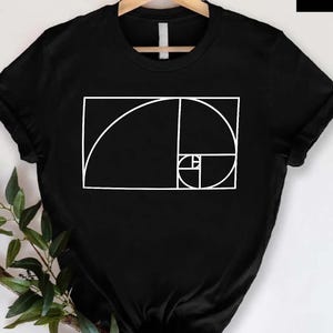 May include: Black t-shirt with a white geometric design featuring a spiral and rectangular shapes. The shirt is hanging on a wooden hanger. A gold logo with the text "De LUX" is in the upper right corner.