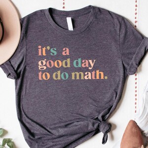 May include: Dark gray t-shirt with the text "it's a good day to do math" in colorful letters. The shirt is styled with light-wash jeans, brown flats, and a beige hat. A beaded necklace and sprig of greenery complete the look.