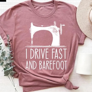May include: A dusty rose t-shirt featuring a white graphic of a sewing machine and the text "I DRIVE FAST AND BAREFOOT". The shirt is styled with a rolled sleeve and a knotted hem. A neutral-toned hat and white mug are also in the image.