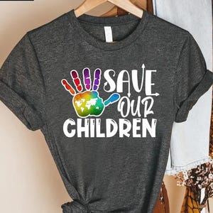 Peut inclure: T-shirt gris fonc&eacute; avec l'inscription blanche "SAVE OUR CHILDREN". Le motif comprend une empreinte de main color&eacute;e avec un globe dans la paume. Le t-shirt est suspendu &agrave; un cintre en bois.