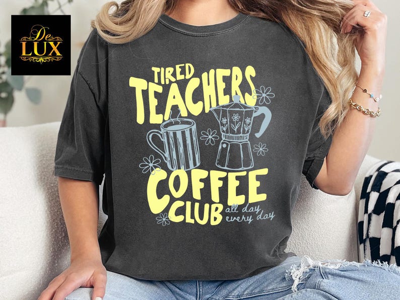 K&ouml;nnte beinhalten: Dunkelgraues T-Shirt mit gelber und hellblauer Grafik. Die Grafik zeigt "Tired Teachers Coffee Club" mit Illustrationen einer Kaffeetasse und eines Espressokochers. Der Text "all day, every day" ist ebenfalls enthalten.