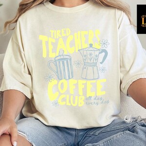 K&ouml;nnte beinhalten: Cremefarbenes T-Shirt mit dem Text "Tired Teachers Coffee Club" in Gelb und Hellblau. Das Design umfasst eine Kaffeekanne, eine Tasse und florale Akzente. Das Shirt ist ein l&auml;ssiger, bequemer Stil.