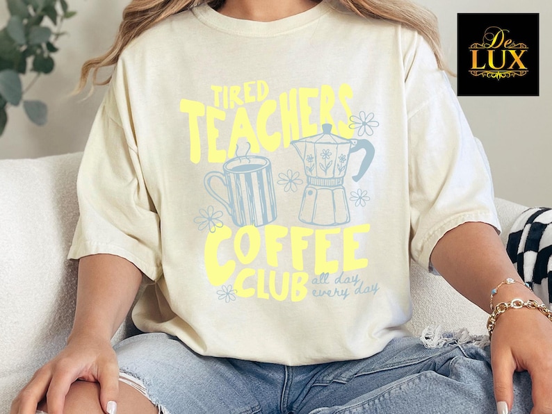 K&ouml;nnte beinhalten: Cremefarbenes T-Shirt mit dem Text "Tired Teachers Coffee Club" in Gelb und Hellblau. Das Design umfasst eine Kaffeekanne, eine Tasse und florale Akzente. Das Shirt ist ein l&auml;ssiger, bequemer Stil.