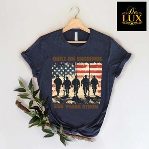 May include: Navy blue t-shirt with a distressed American flag design, soldier silhouettes, and the text "BUILT ON SACRIFICE" and "250 YEARS STRONG". The shirt is displayed on a wooden hanger.