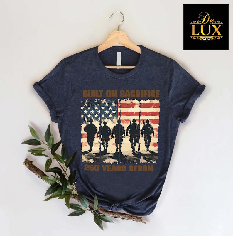 May include: Navy blue t-shirt with a distressed American flag design, soldier silhouettes, and the text "BUILT ON SACRIFICE" and "250 YEARS STRONG". The shirt is displayed on a wooden hanger.