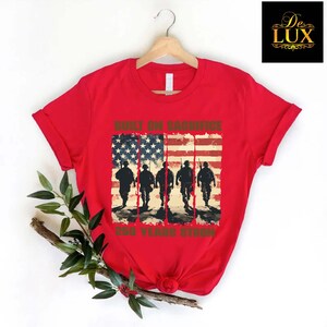 May include: A red t-shirt with a design featuring the American flag and silhouettes of soldiers. The text on the shirt reads "BUILT ON SACRIFICE" and "250 YEARS STRONG". The t-shirt is displayed on a wooden hanger.