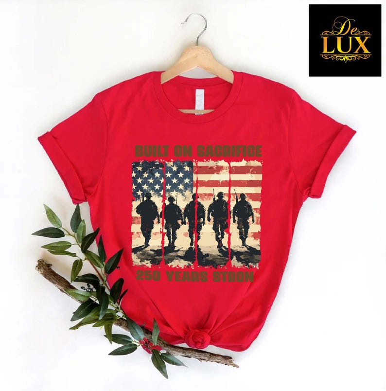 May include: A red t-shirt with a design featuring the American flag and silhouettes of soldiers. The text on the shirt reads "BUILT ON SACRIFICE" and "250 YEARS STRONG". The t-shirt is displayed on a wooden hanger.