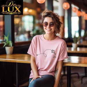 Pu&ograve; includere: Una maglietta rosa chiaro con una grafica di aereo nera e la scritta "Custom Location". La maglietta ha maniche corte. Il logo De Lux &egrave; nell'angolo in alto a sinistra. La persona indossa occhiali da sole.