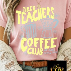 K&ouml;nnte beinhalten: Rosafarbenes T-Shirt mit dem Text "Tired Teachers Coffee Club" in Gelb, mit hellblauen Kaffee-Grafiken. Der Satz "all day every day" steht unter dem Club-Text. Ein brauner G&uuml;rtel und blaue Jeans sind sichtbar.