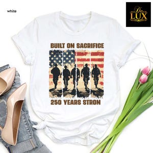 May include: White t-shirt with a distressed American flag design and soldier silhouettes. The text reads "BUILT ON SACRIFICE" above the flag and "250 YEARS STRON" below. The shirt has a crew neck and is made of a soft material.