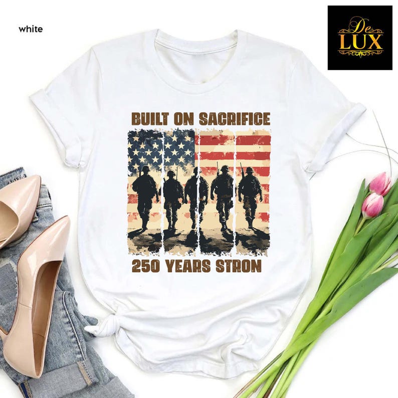 May include: White t-shirt with a distressed American flag design and soldier silhouettes. The text reads "BUILT ON SACRIFICE" above the flag and "250 YEARS STRON" below. The shirt has a crew neck and is made of a soft material.