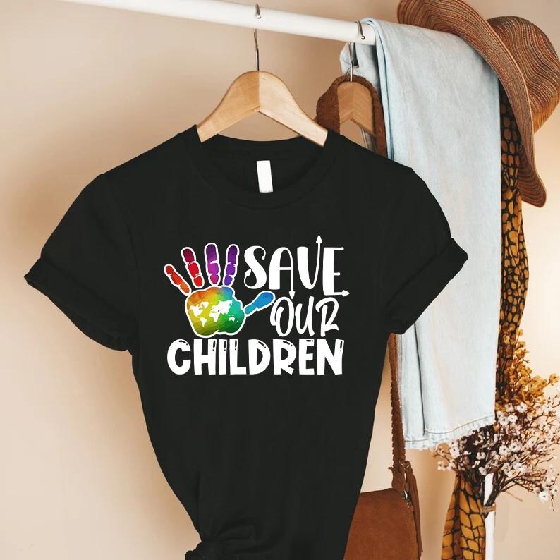 Peut inclure: T-shirt noir avec l'inscription blanche "SAVE OUR CHILDREN". Le motif comprend une empreinte de main color&eacute;e avec une carte du monde dans la paume. Le t-shirt est suspendu &agrave; un cintre en bois.