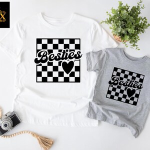 May include: Two t-shirts, one white and one gray, featuring a black and white checkered design with the word "Besties" and a heart. The white shirt is adult size, and the gray shirt is smaller, likely for a child.