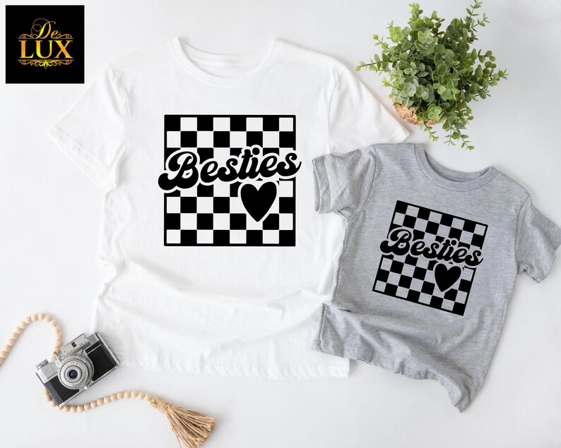 May include: Two t-shirts, one white and one gray, featuring a black and white checkered design with the word "Besties" and a heart. The white shirt is adult size, and the gray shirt is smaller, likely for a child.