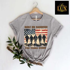 May include: A grey t-shirt with a distressed American flag design and soldier silhouettes. The text "BUILT ON SACRIFICE" and "250 YEARS STRONG" are printed above and below the flag graphic. A De Lux logo is in the upper right corner.